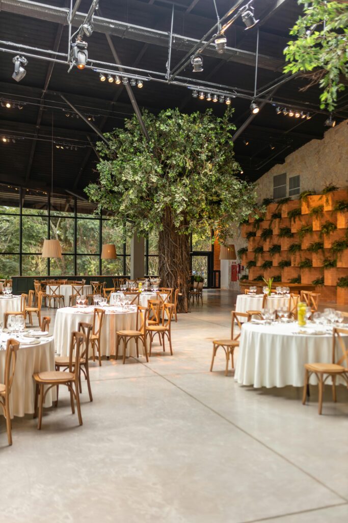 Chic dining area in a hall with elegant table settings surrounded by greenery and trees.