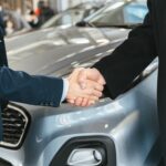 A close-up of two people shaking hands in front of a car in an indoor setting.